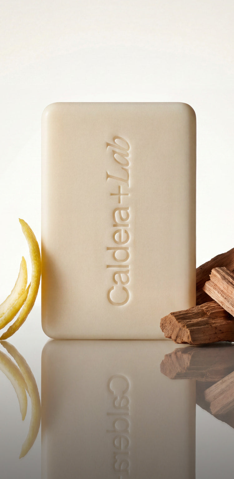 Bar of soap with 'Caldera Lab' branding on a reflective surface with lemon and cinnamon sticks.