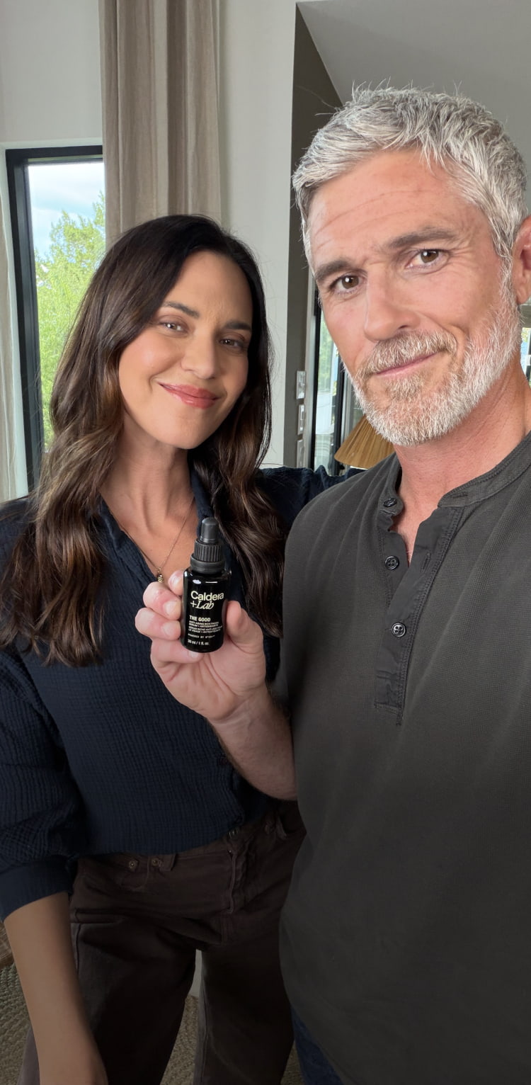 A man and a woman taking a selfie indoors,  holding a bottle of The Good.