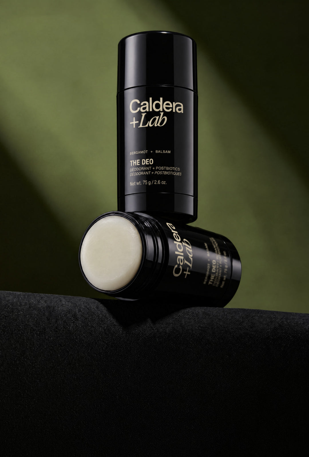Two black Caldera + Lab deodorant sticks on a dark surface against an olive green background, with one stick standing upright and the other lying on its side showing the cream-colored deodorant.