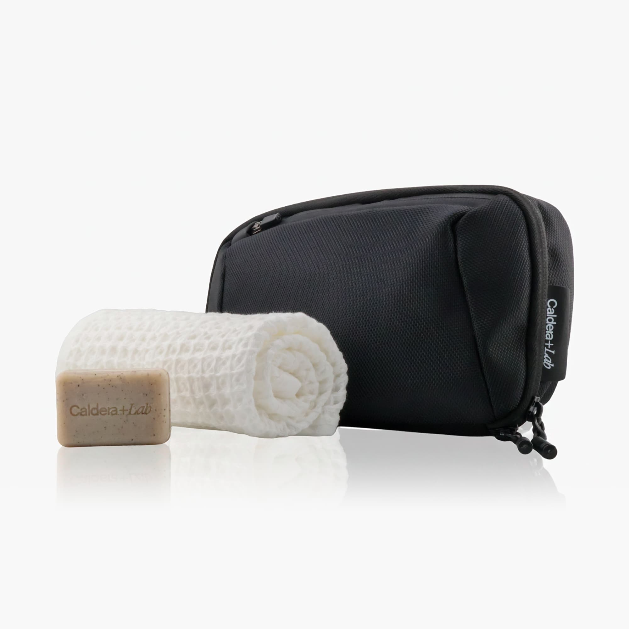 Black toiletry bag with a white washcloth and bar of soap on a light gray background